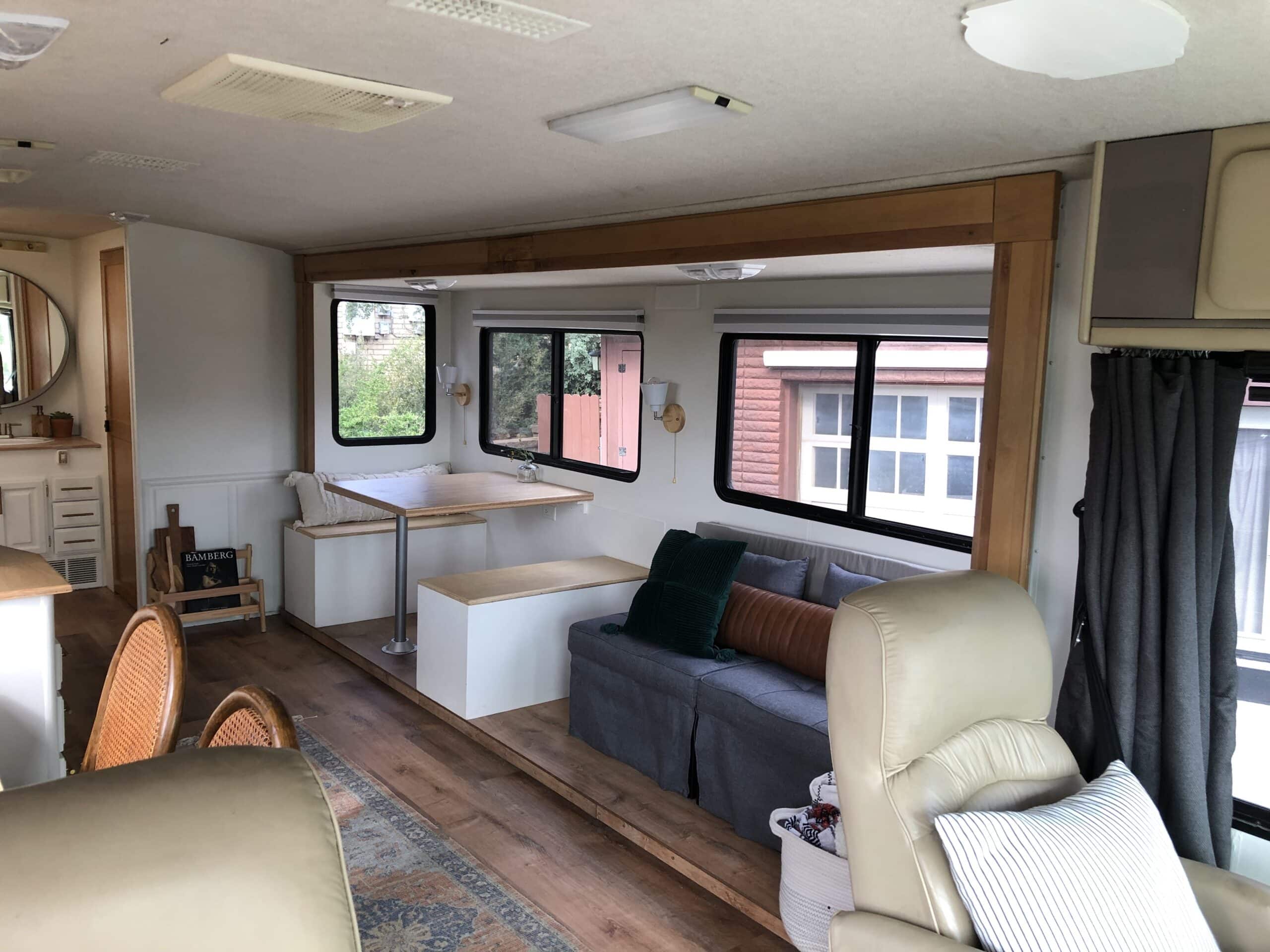 RV interior showing dinette, sofa, and recliner with large windows.